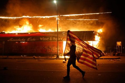A protester runs in front of a burning building in Minneapolis waving an upside down flag, a symbol of emergency or danger. 